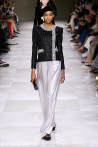 Model walks the runway in a black sequined jacket with silver trim and matching silver satin wide-leg pants, holding a small clutch.
