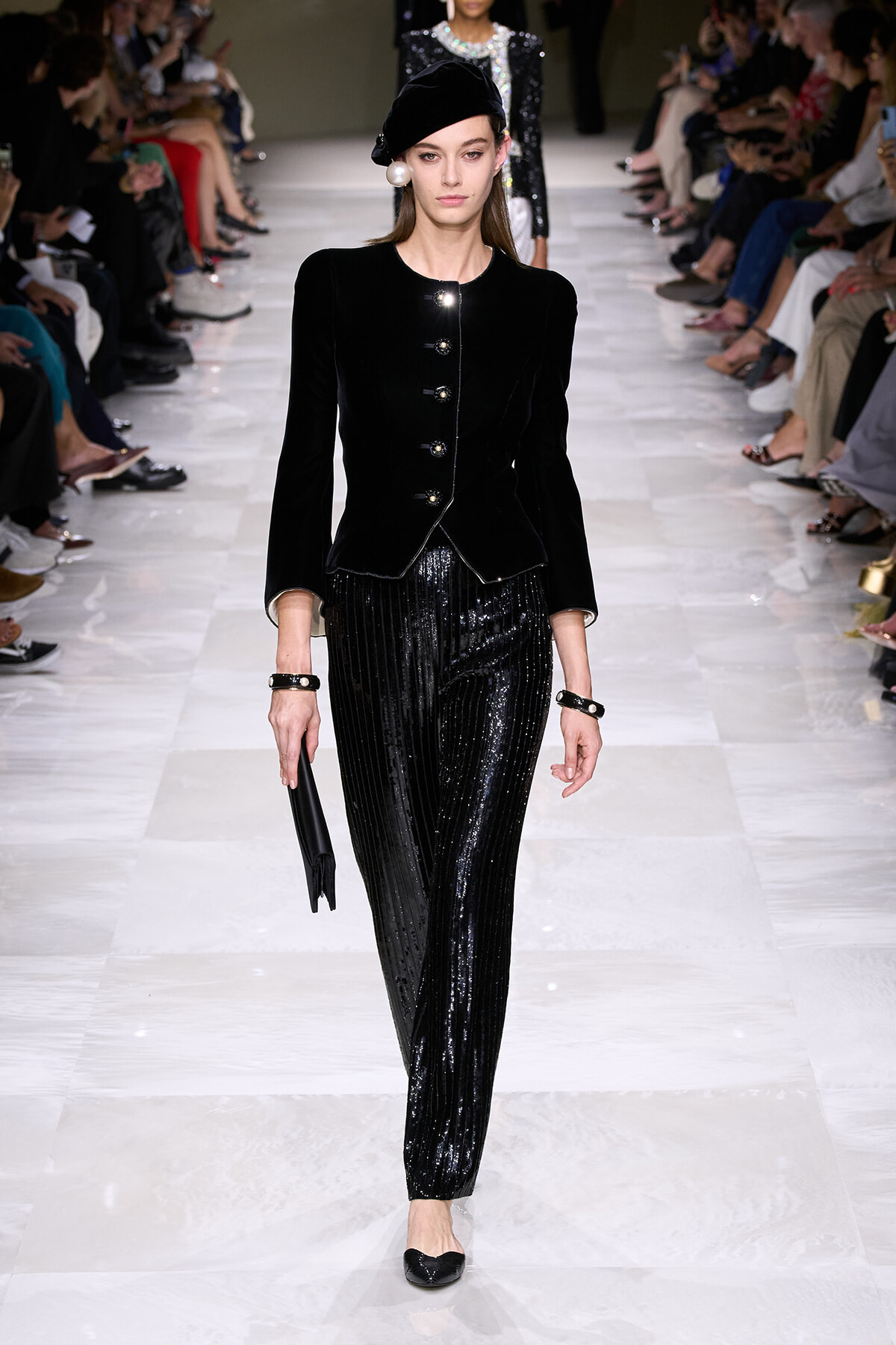 Model on a runway wearing a black velvet buttoned jacket and sparkly black pants, with a beret and pearl earrings and holding a black clutch.