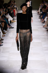 Model walks the runway in a black velvet top and glittering, sheer-sequin pants, carrying a black clutch; audience rows line the catwalk.