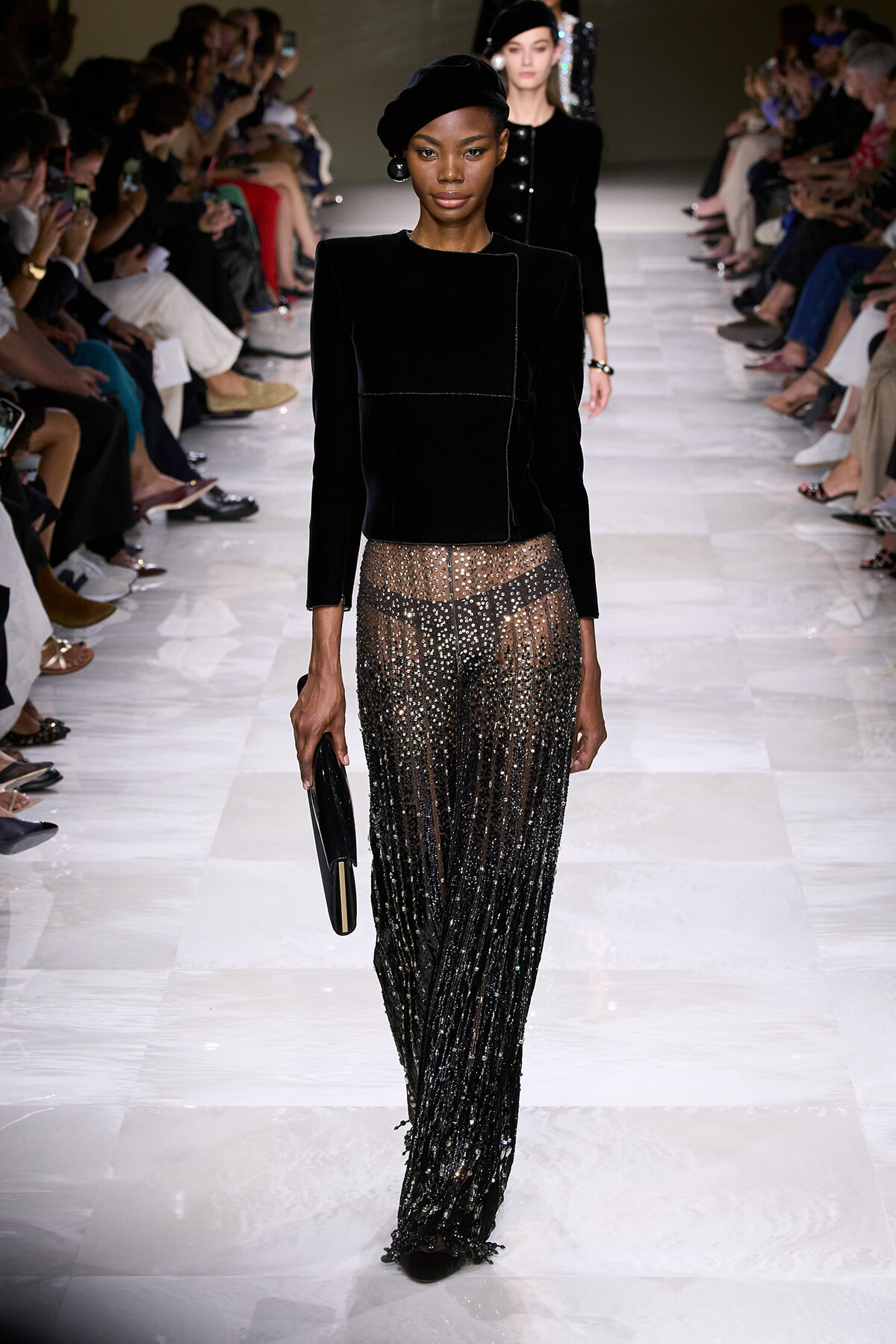Model walks the runway in a black velvet top and glittering, sheer-sequin pants, carrying a black clutch; audience rows line the catwalk.