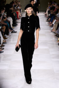 Model walks the runway in a black velvet jumpsuit with large white buttons and a matching beret, holding a small black clutch.