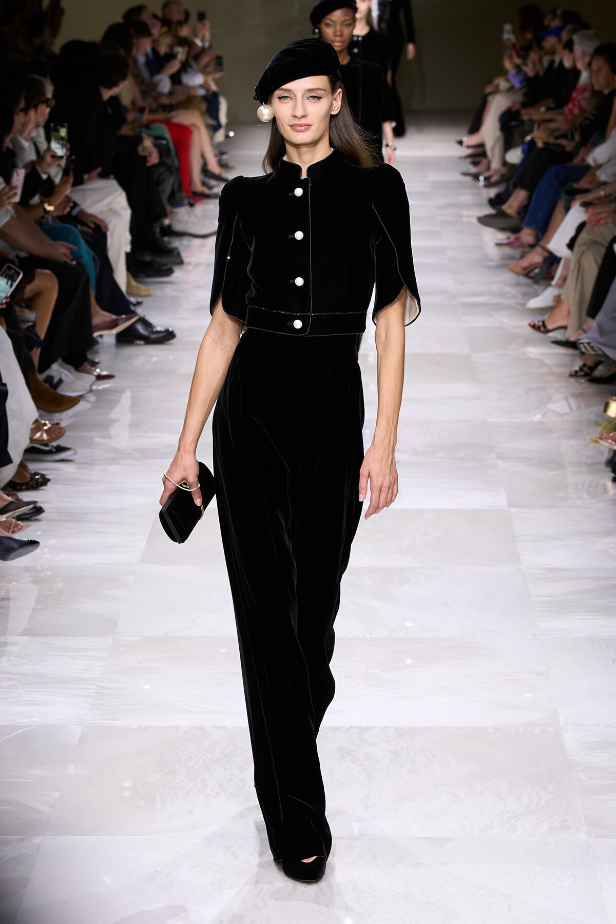 Model walks the runway in a black velvet jumpsuit with large white buttons and a matching beret, holding a small black clutch.