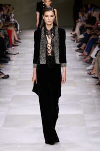 Model walks the runway in a black velvet suit with a silver embellished scarf and multiple layered black beaded necklaces.