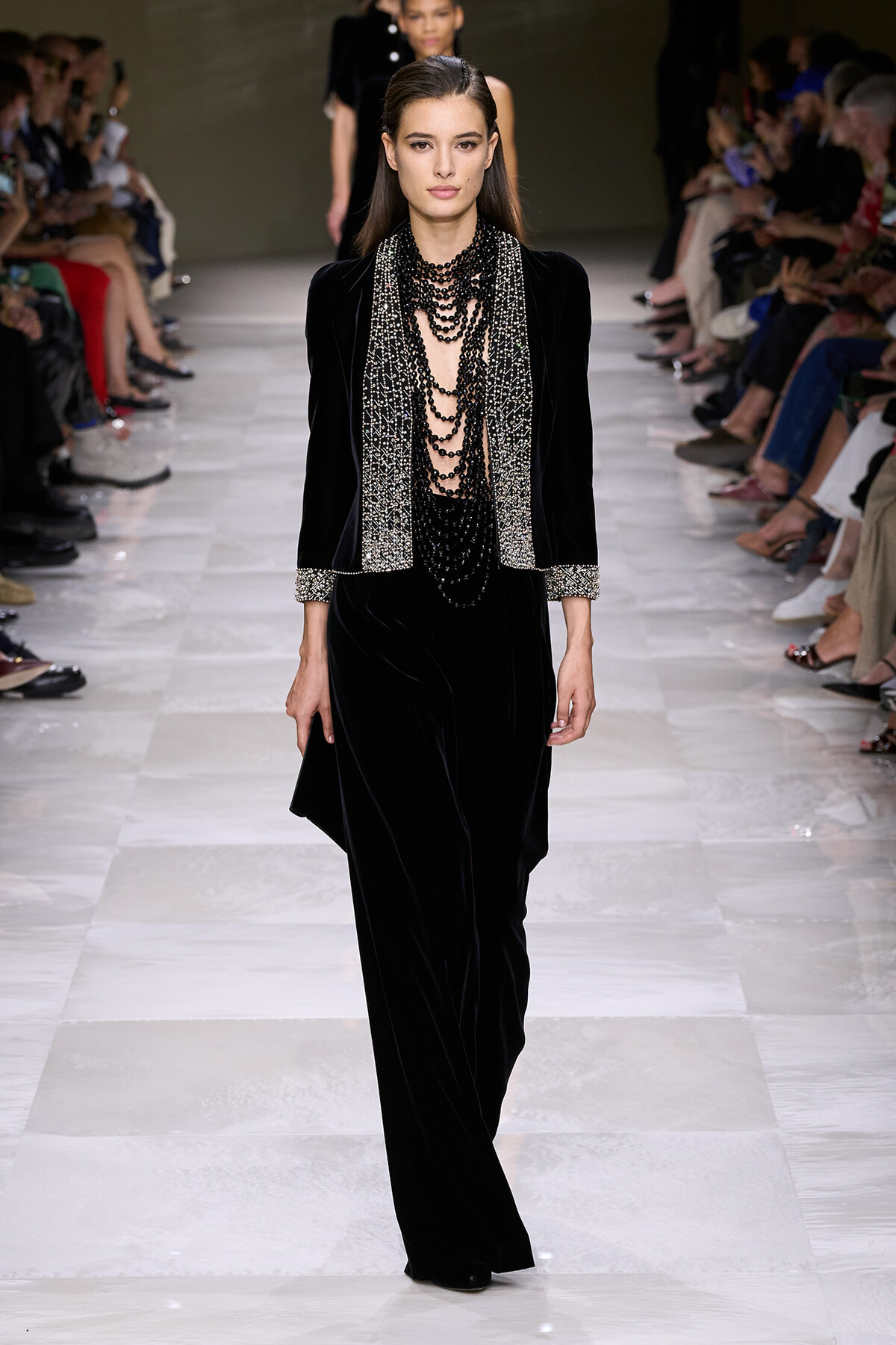 Model walks the runway in a black velvet suit with a silver embellished scarf and multiple layered black beaded necklaces.