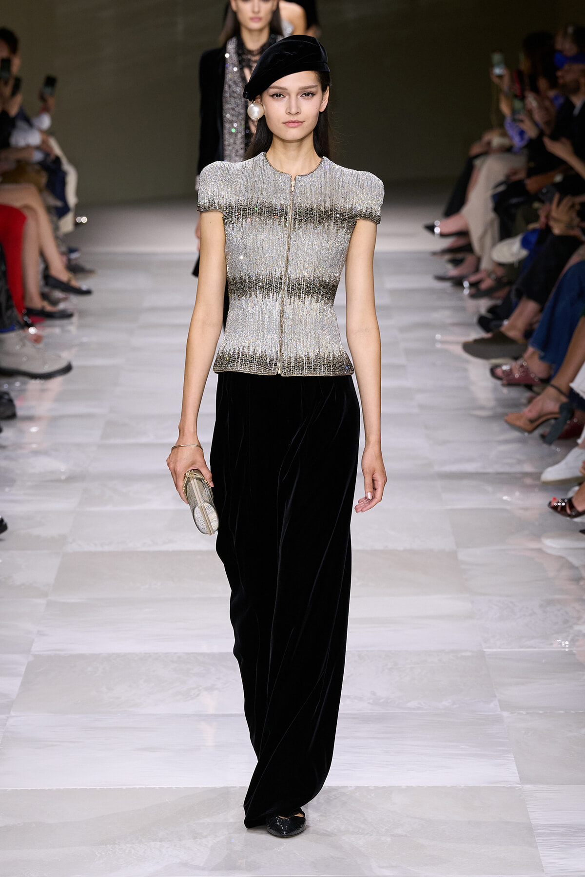 Runway model wearing a shimmering silver beaded top with short sleeves, black velvet wide-leg pants, a black beret, large pearl earring, and a metallic clutch.