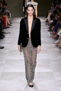 Model walking a runway in a black velvet blazer with an open front revealing a pearl necklace and sheer, beaded pants, carrying a small clutch with a chain strap.
