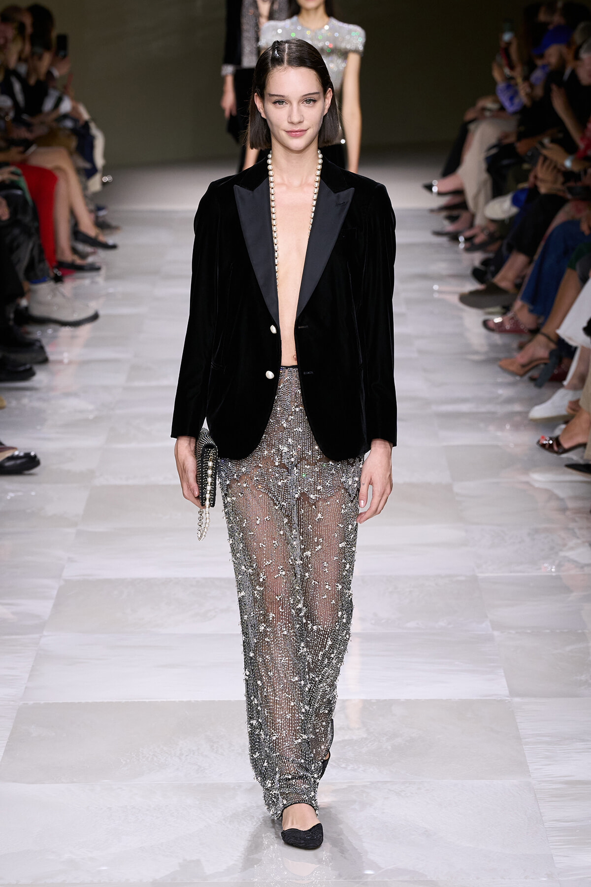 Model walking a runway in a black velvet blazer with an open front revealing a pearl necklace and sheer, beaded pants, carrying a small clutch with a chain strap.
