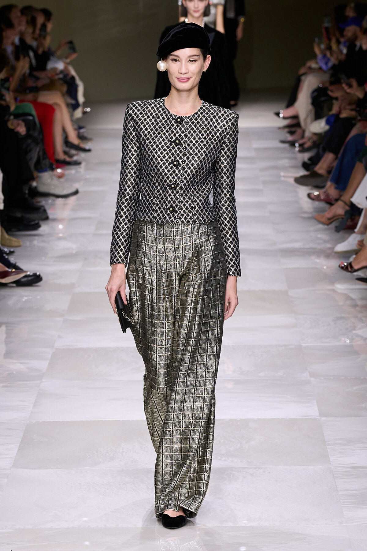 Model walking the runway in a black-and-white patterned jacket with gold buttons and metallic plaid wide-leg pants, wearing a black beret and large pearl earring.