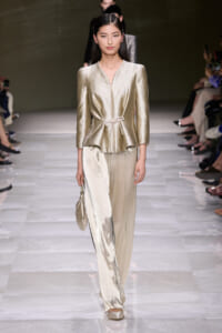 Model walks the runway in a gold metallic blouse with a matching wide-leg satin pant and small gold handbag on her left side.