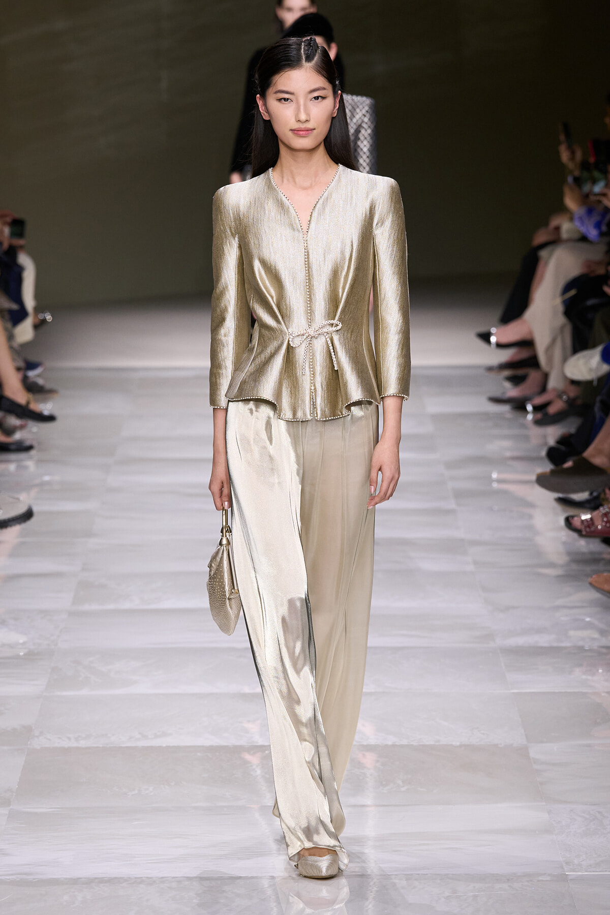 Model walks the runway in a gold metallic blouse with a matching wide-leg satin pant and small gold handbag on her left side.