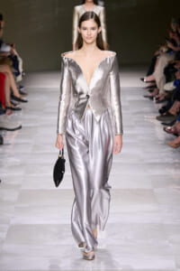 Model walks the runway in a metallic silver off-shoulder blazer with a deep V neckline and peplum, paired with silver wide-leg satin pants and a small black handbag.
