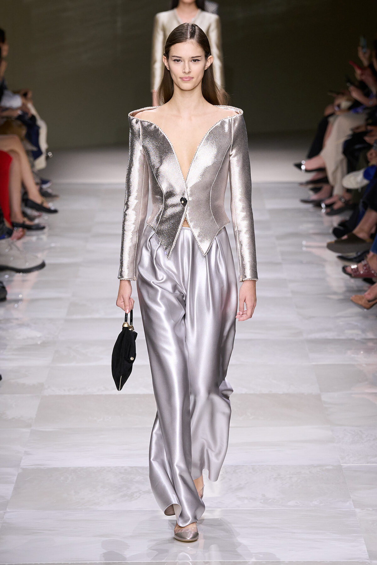 Model walks the runway in a metallic silver off-shoulder blazer with a deep V neckline and peplum, paired with silver wide-leg satin pants and a small black handbag.