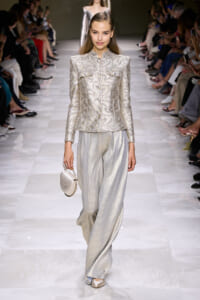 Model walking a fashion runway in a silver embroidered suit with a structured jacket and wide-leg pants, carrying a small silver handbag.