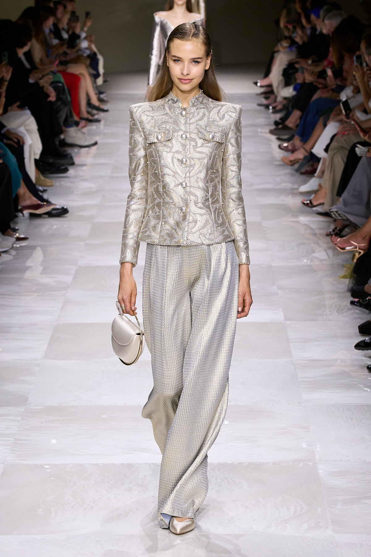 Model walking a fashion runway in a silver embroidered suit with a structured jacket and wide-leg pants, carrying a small silver handbag.