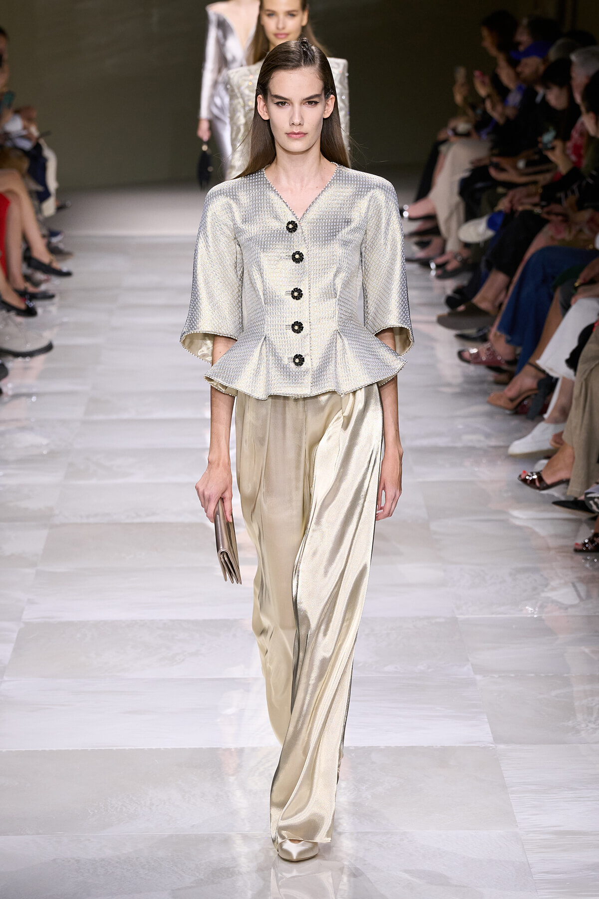 Model walks the runway in a silver textured peplum top with black buttons and wide ivory satin pants, holding a beige clutch