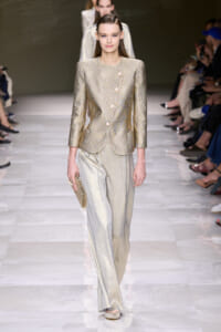 Female model walks the runway wearing a metallic champagne-gold textured blazer with matching wide-leg pants, holding a beige clutch.
