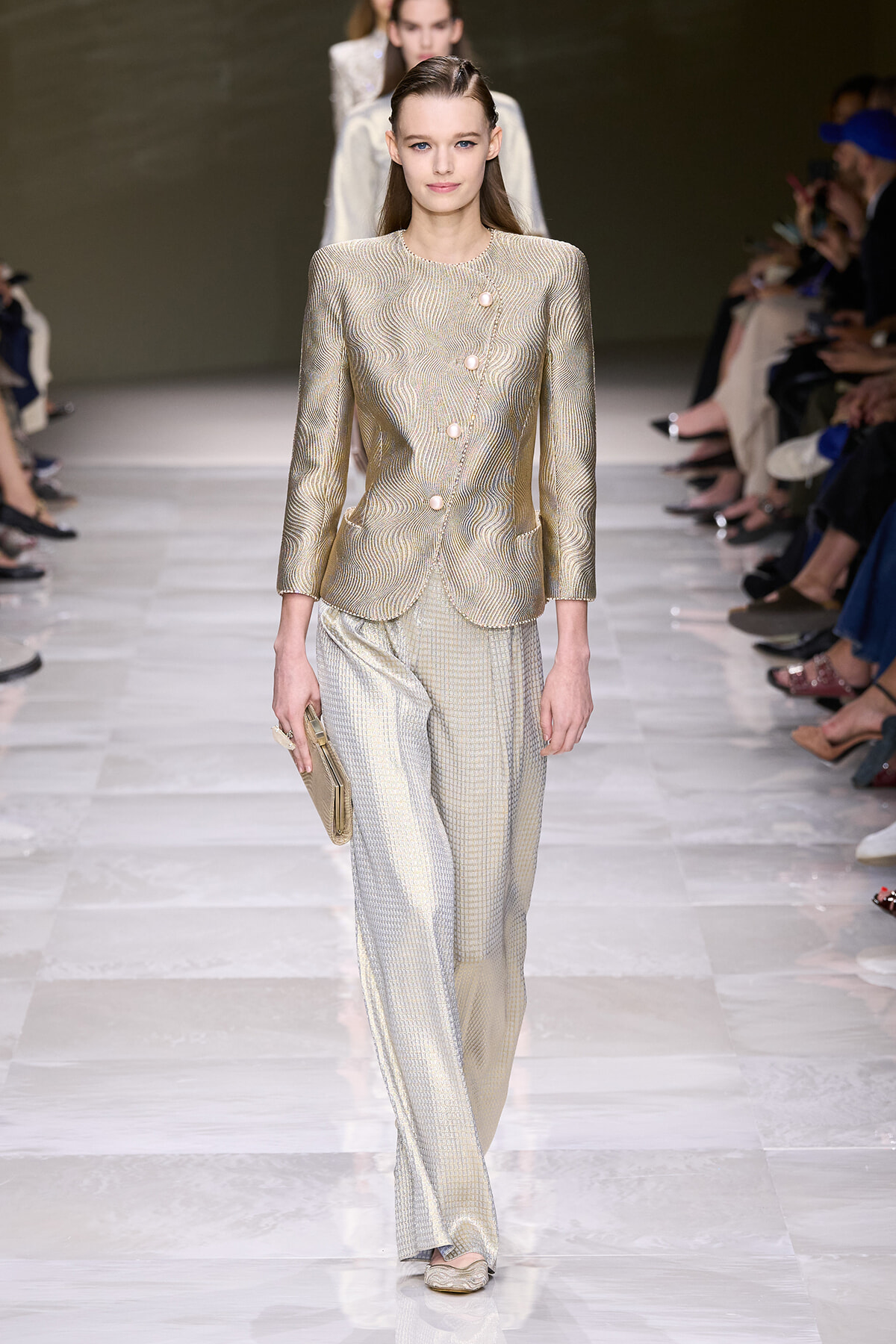 Female model walks the runway wearing a metallic champagne-gold textured blazer with matching wide-leg pants, holding a beige clutch.