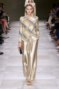 Model walking a runway in a beige patterned long-sleeve top and gold satin wide-leg pants, holding a small clutch.