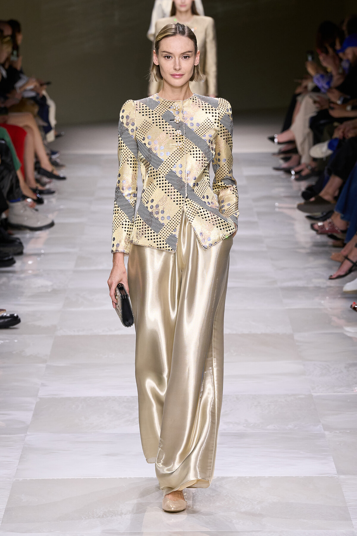 Model walking a runway in a beige patterned long-sleeve top and gold satin wide-leg pants, holding a small clutch.