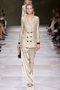 Model walks the runway in a cream textured double-breasted blazer with black buttons and satin wide-leg trousers, clutch in hand.