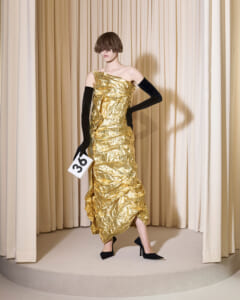 Model in a crinkled gold one-shoulder gown with long black gloves, holding a white card labeled 36, posed on a beige-curtain backdrop.