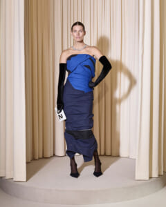Model in a blue strapless color‑blocked gown with long black opera gloves and a chunky necklace, standing on a circular runway holding number 32.