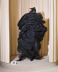 Model wearing a dramatic, oversized black sculptural gown made of shiny fabric on a circular runway; number 39 card visible.
