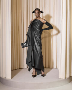 Model in a black one-shoulder leather gown with a draped, asymmetrical skirt stands on a circular runway, holding a black clutch with '35' on it.