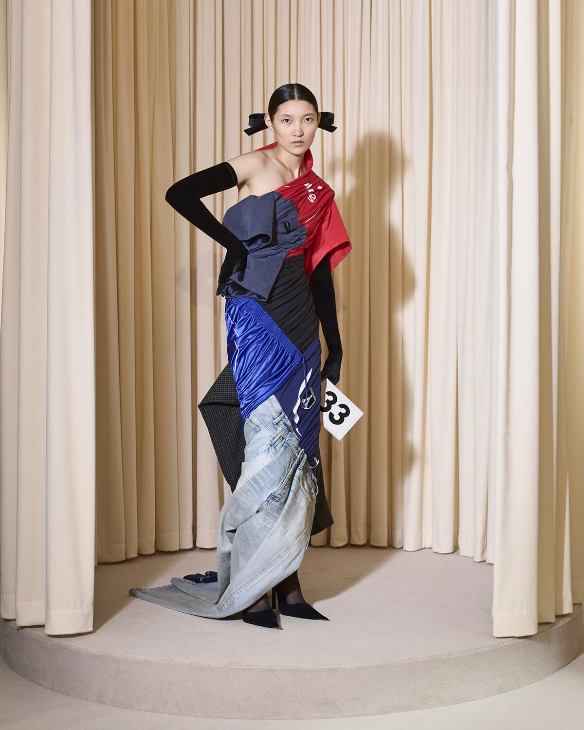 Model posing on a circular runway wearing layered red, black, blue fabric with a long denim fold; number 33 tag held in hand, beige stage curtains behind.