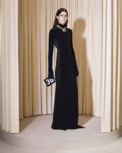 Model wearing a long black high-neck gown with a jeweled necklace, standing before cream curtains, holding a small black clutch with '30' on it.