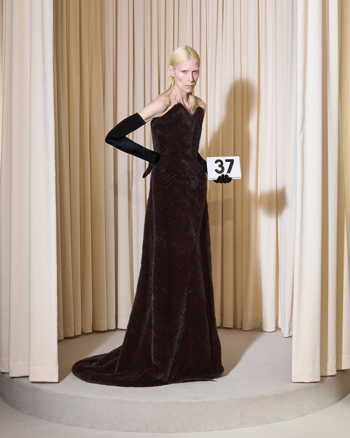 Model in a floor-length black fur gown and long black gloves stands on a circular stage with beige curtains, holding a card that reads 37.