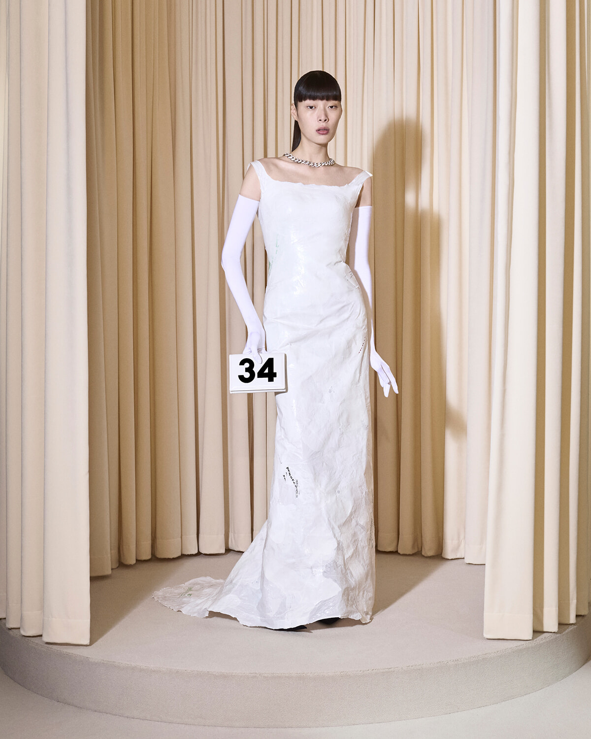 Model in a white off-shoulder gown with long white gloves stands on a circular runway holding a sign that reads 34.
