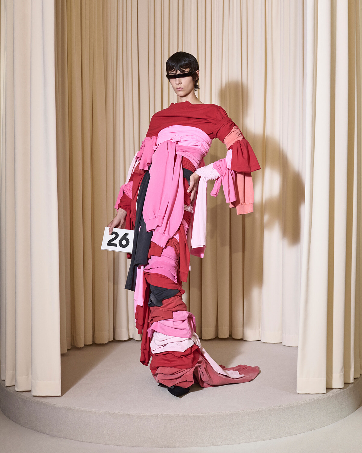 Model on a runway wearing a red top with layered pink fabric wrapped around her, holding a card with the number 26, in front of beige curtains on a circular stage.