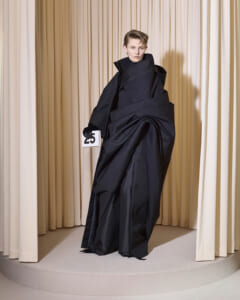 Model in an oversized black coat on a circular runway, holding a card numbered 25, beige curtain backdrop.