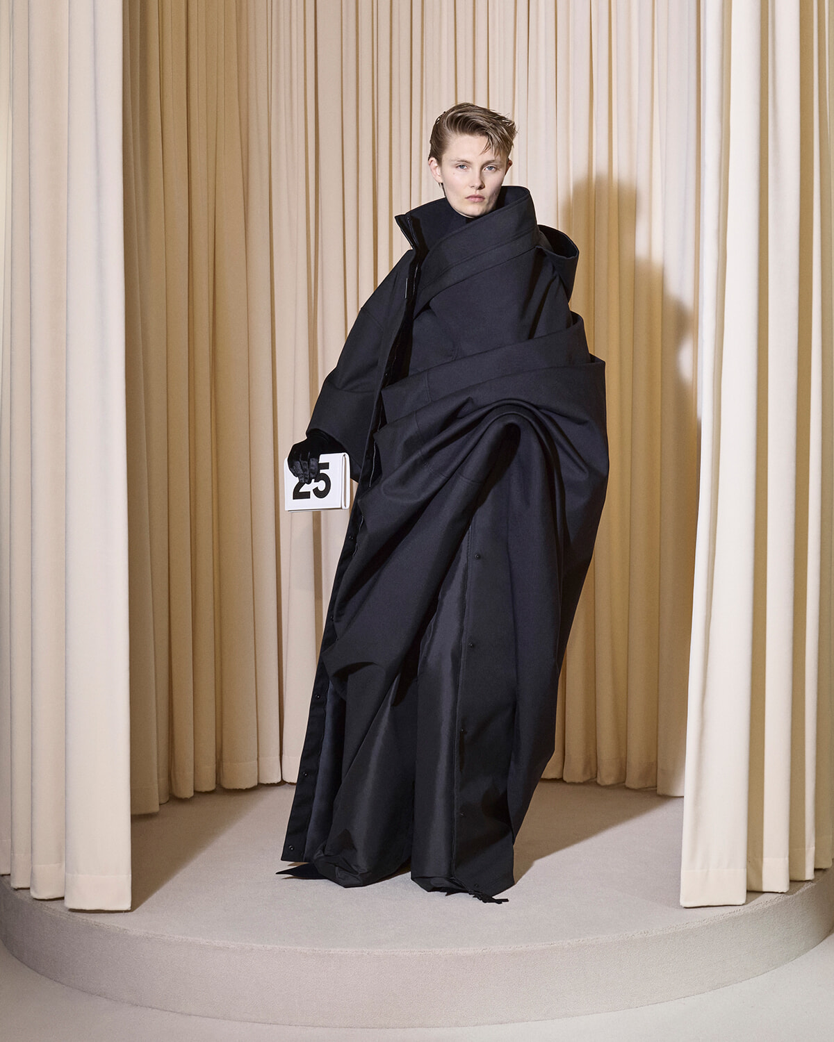 Model in an oversized black coat on a circular runway, holding a card numbered 25, beige curtain backdrop.