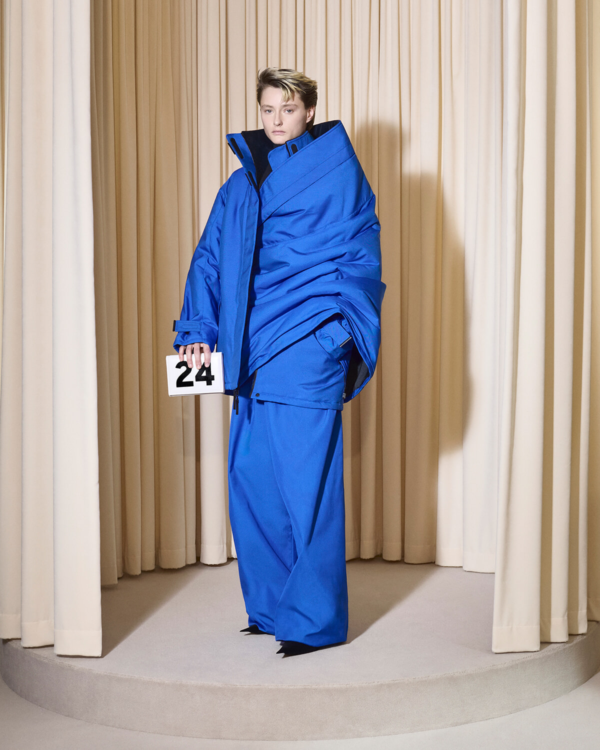 Model in an oversized cobalt blue padded coat and wide pants on a beige curtain backdrop, holding a white card reading 24 in a fashion show setting.