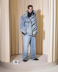 Male model in an oversized light-wash denim jacket and wide-leg jeans stands on a circular pedestal against beige curtains; a '23' sign is in front.