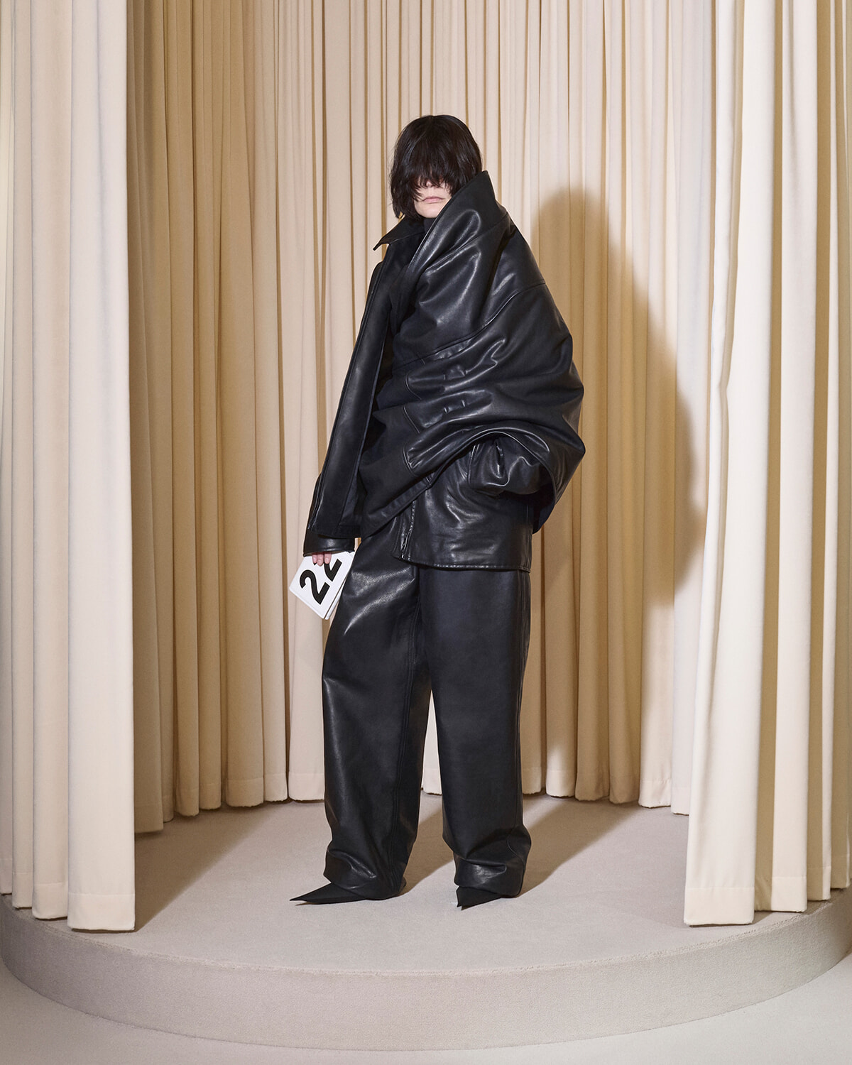 Model in an oversized black leather ensemble on a circular stage, holding a numbered card (2) against beige curtain backdrop.
