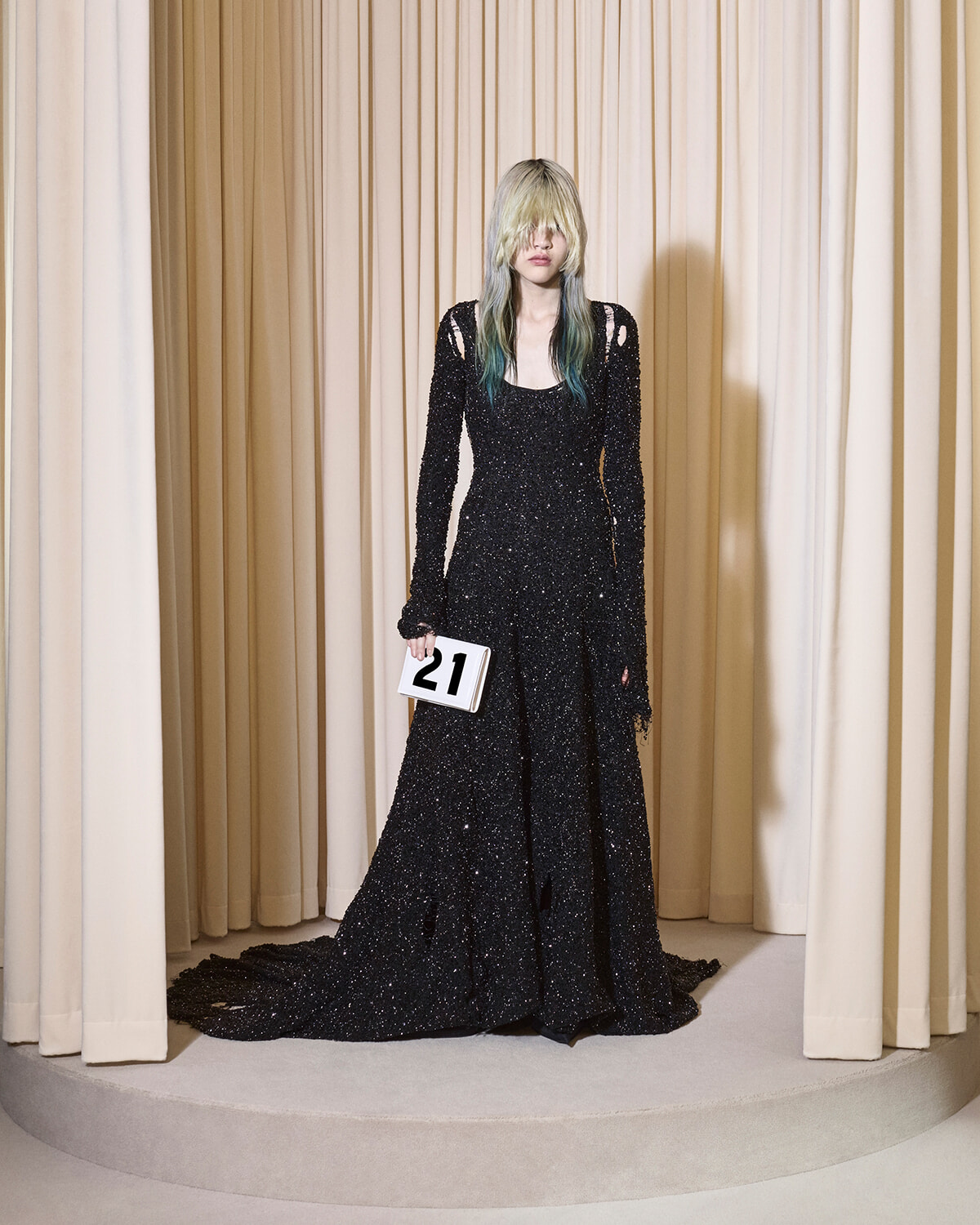 Model in a glittery black gown with blue-tinted hair, standing on a circular platform while holding a white clutch marked 21.