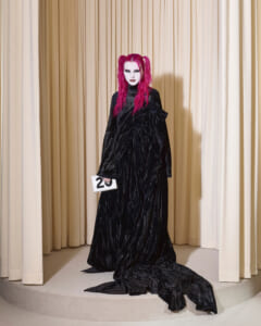 Person with bright pink hair in a floor-length black velvet gown stands on a circular stage, holding a white card marked '20' against beige curtains.