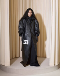 Model in an oversized black leather coat and gloves stands on a circular beige platform, holding a white number 19 tag.