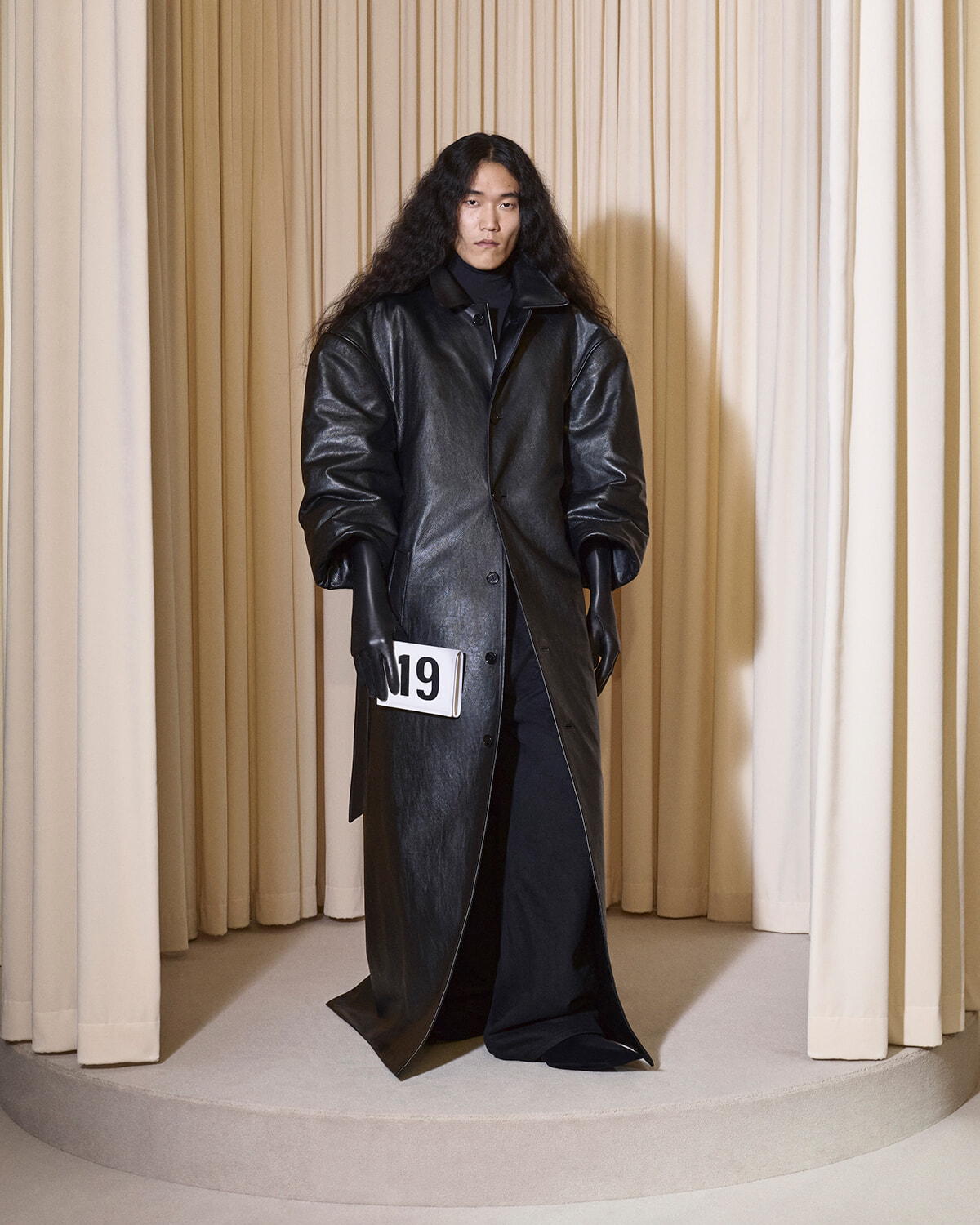 Model in an oversized black leather coat and gloves stands on a circular beige platform, holding a white number 19 tag.