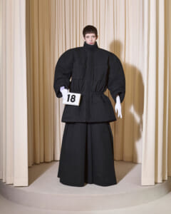 Model wearing an oversized black jacket and wide black pants on a runway, holding a white sign with the number 18, beige curtain backdrop.