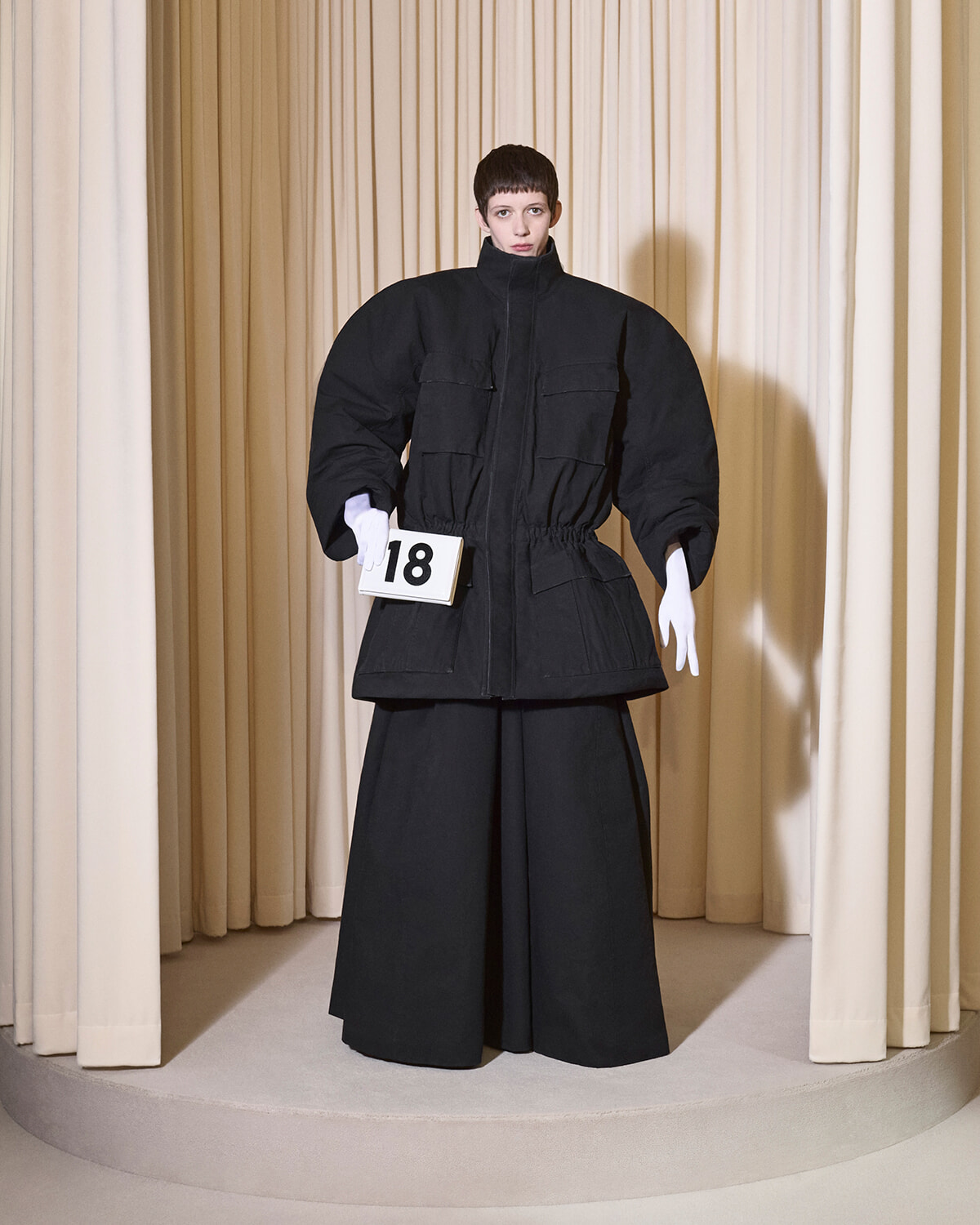 Model wearing an oversized black jacket and wide black pants on a runway, holding a white sign with the number 18, beige curtain backdrop.