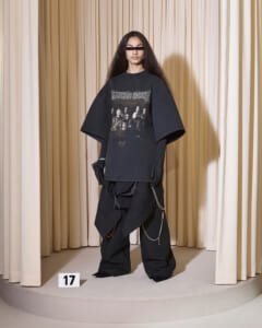 Model wearing an oversized black graphic T-shirt and baggy pants with chains, posed on a circular runway with beige curtains behind.