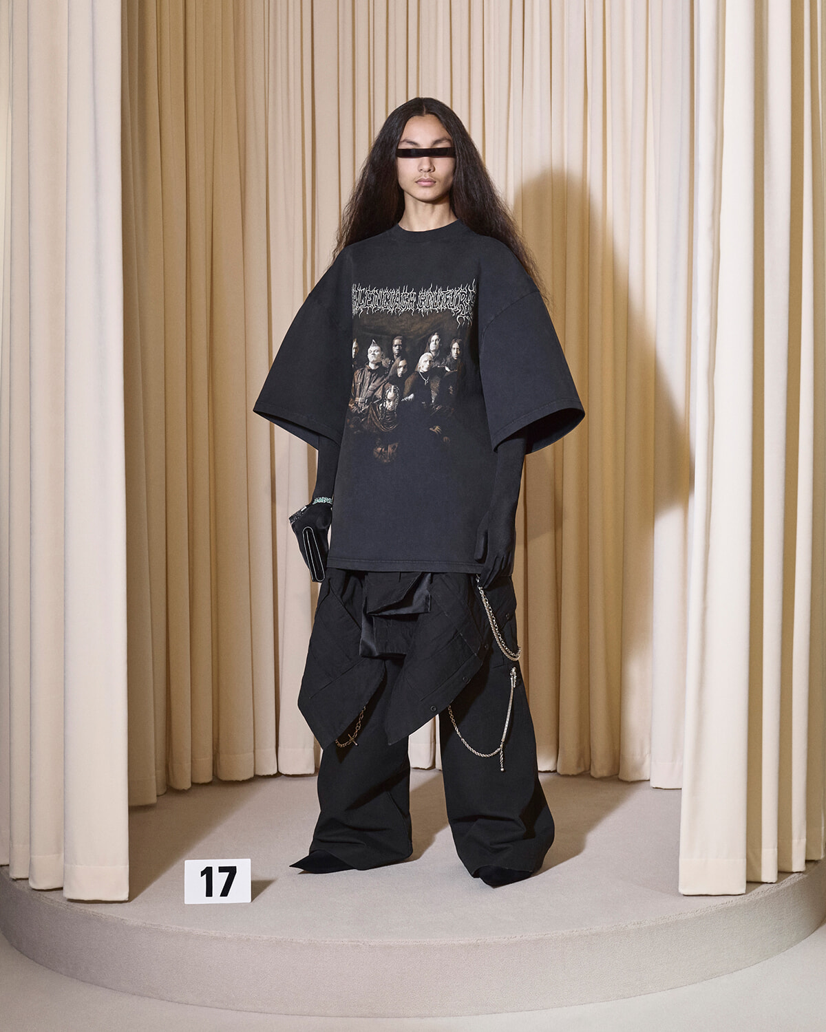 Model wearing an oversized black graphic T-shirt and baggy pants with chains, posed on a circular runway with beige curtains behind.