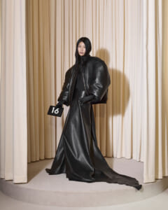 Model in an oversized black leather coat and long skirt on a circular runway, holding a black '16' placard against cream curtains.