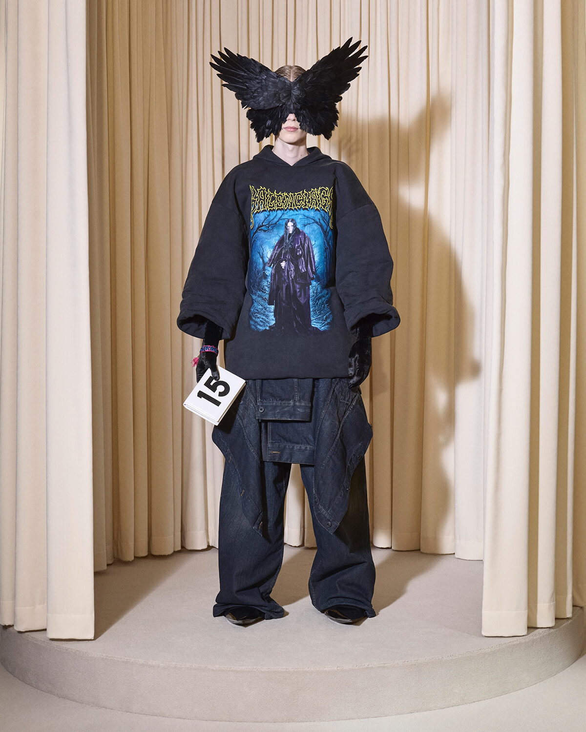 Model on a runway wearing an oversized black hoodie with a blue graphic, paired with baggy cargo pants and a large black feather mask obscuring the face, holding a 15 tag plate.