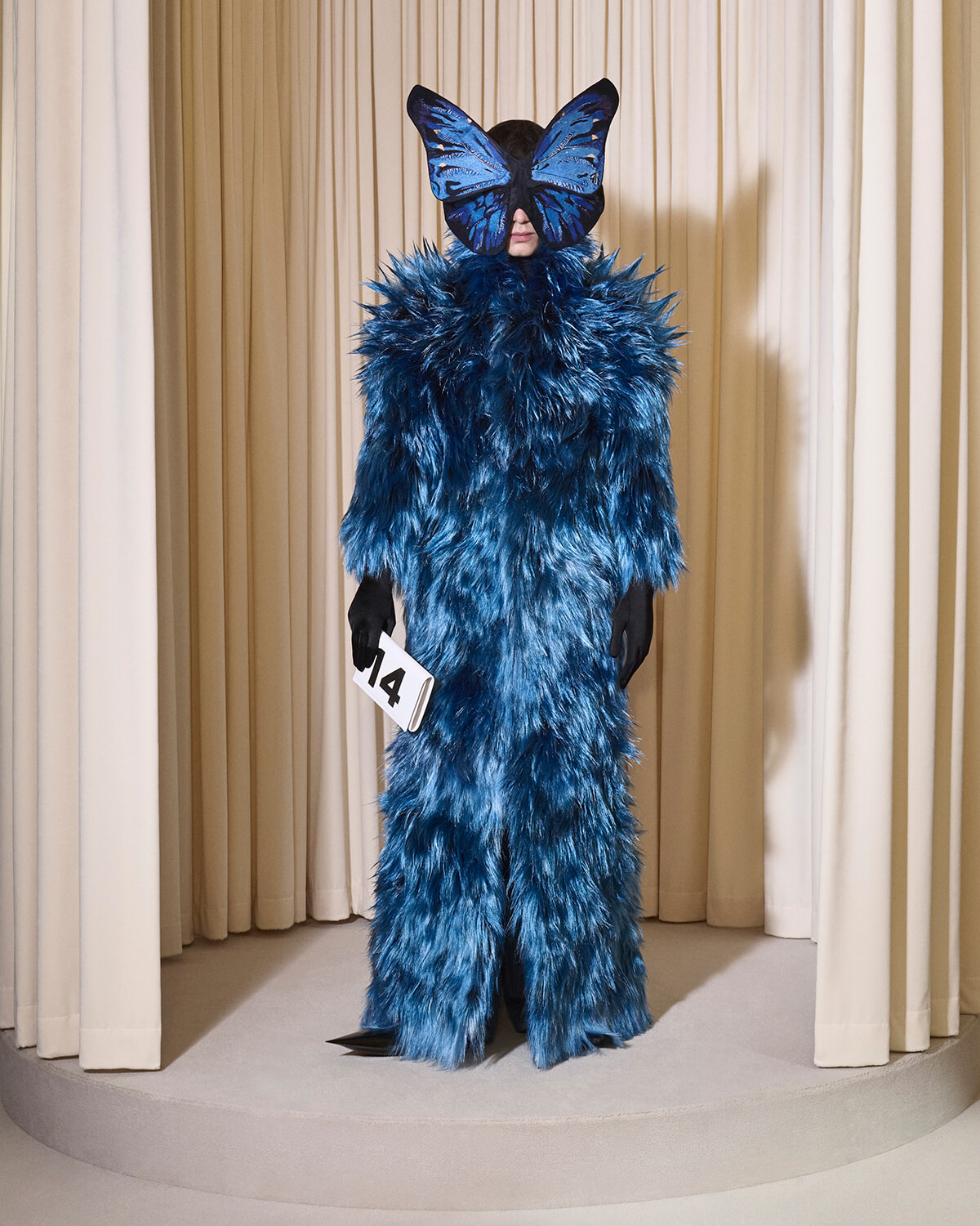 Person in a blue feathered costume with an oversized blue butterfly mask, standing on a round platform.