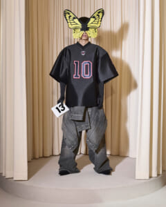Person on a stage wearing a black jersey with the number 10, oversized gray pants, a large yellow butterfly mask, and holding a card with the number 13.